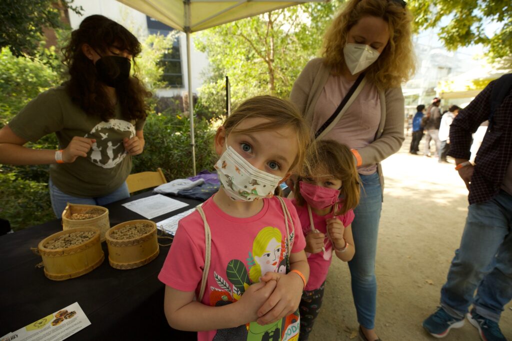 Photo of a young child with blond hair and pale skin wearing a face mask looking up curiously at the photographer. Behind her are more people, and a table with woven baskets.