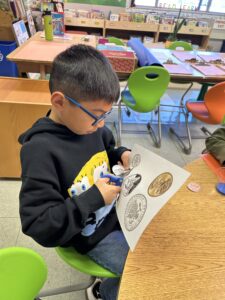 A photo of a child with light brown skin and short, straight, black hair sitting in a classroom and cutting out images of coins from a piece of paper.