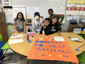 A photo of five kids standing behind a table in a classroom: a child with light brown skin and shoulder-length, straight, black hair; a child with light brown skin and wearing a beanie; a child with light brown skin and short, straight, black hair; a child with light brown skin and long, wavy, black hair; a child with dark skin and short, curly, brown hair. On the table in front of them is a large poster that says, "Donate to help animals / Thank you!"