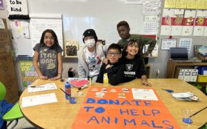 A photo of five kids standing behind a table in a classroom: a child with light brown skin and shoulder-length, straight, black hair; a child with light brown skin and wearing a beanie; a child with light brown skin and short, straight, black hair; a child with light brown skin and long, wavy, black hair; a child with dark skin and short, curly, brown hair. On the table in front of them is a large poster that says, "Donate to help animals / Thank you!"